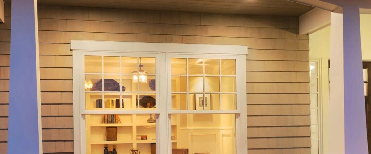 Close-up view of light brown cedar shake siding surrounding white double-hung windows that look into a well-lit living room with bookshelves.