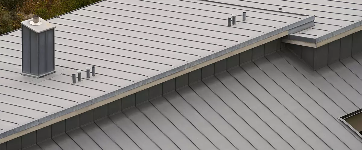 High-angle view of a grey standing seam metal roof featuring a square chimney and ventilation pipes.