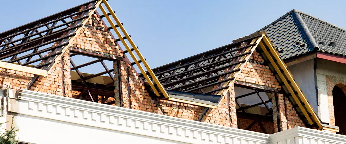 Residential house under construction showing exposed red brick walls and the complex wooden roof truss framework, ready for the next stages of roofing and insulation.