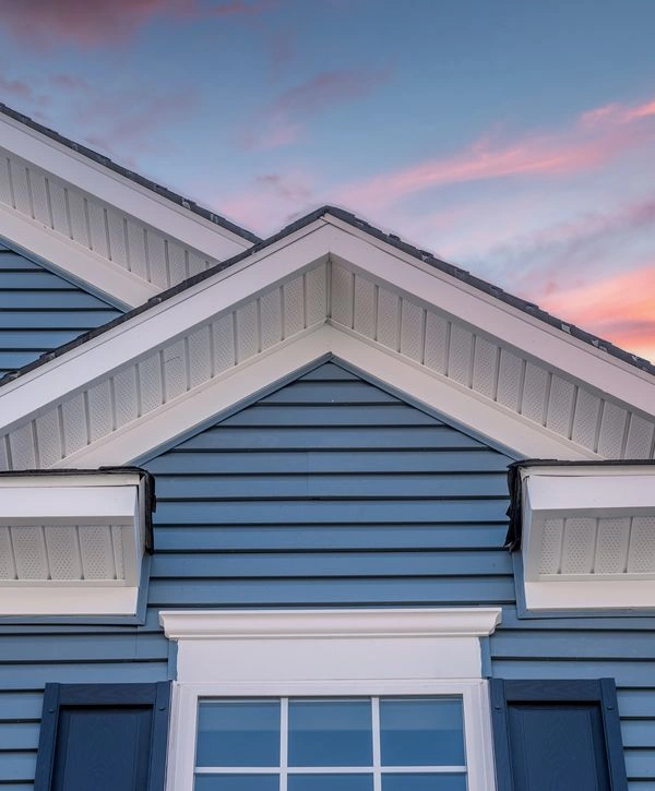 Low-angle view of a home exterior showing blue horizontal siding and white soffit trim during a roof replacement project against a sunset sky.