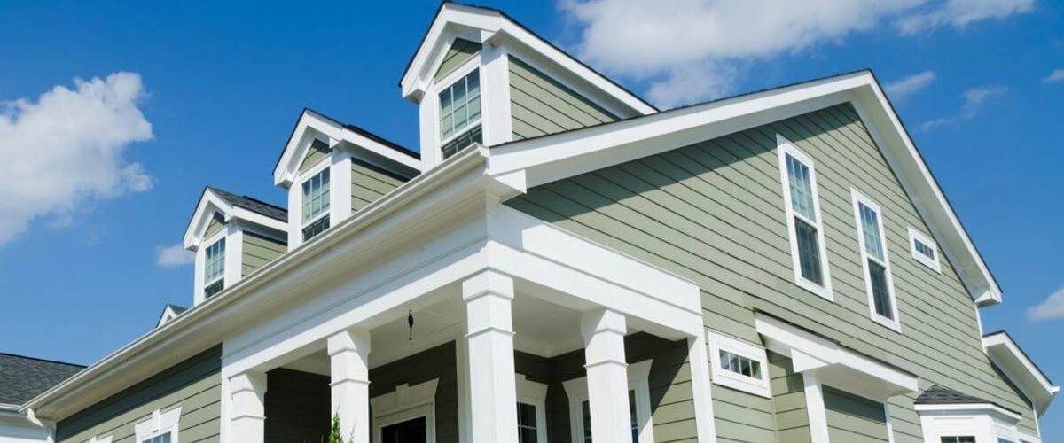 Low angle view of a sage green home exterior featuring white pillars, dormer windows, and clean lap siding lines.