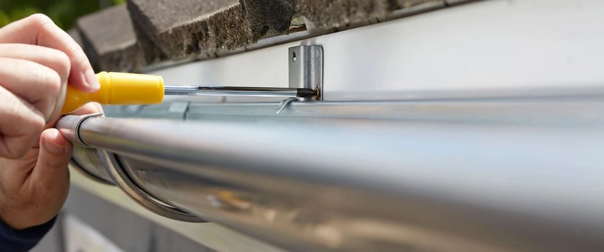 Detailed view of a hand using a yellow-handled screwdriver to secure a metal gutter bracket into the fascia board under a shingled roof.