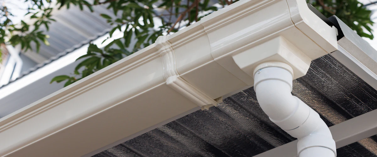 A detailed close-up shot of a clean white vinyl gutter showing the corner joint connection and the attached downspout pipe under the eaves of a building.