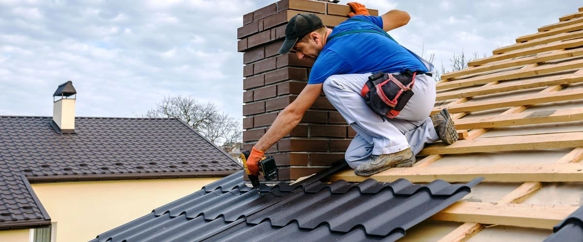 A construction worker crouching on a rooftop using a cordless power drill to secure dark metal roofing panels near a brick chimney structure.