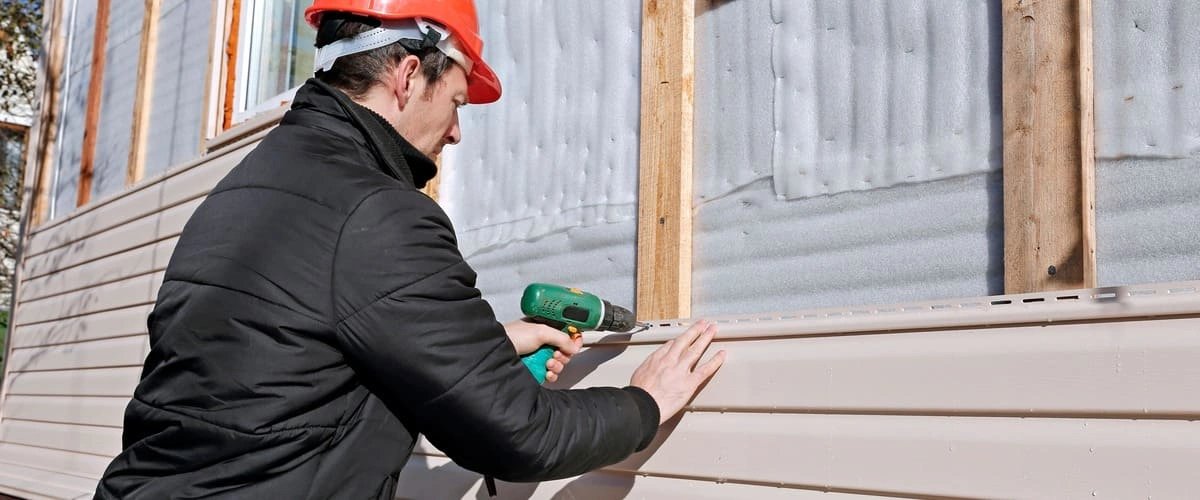 A professional contractor wearing an orange hard hat and black jacket using a power drill to install or repair tan vinyl siding panels on a residential home exterior.