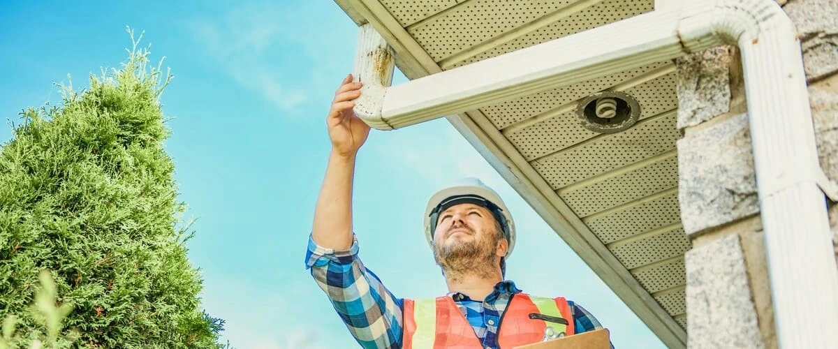 A home inspector in a safety vest and white hard hat examines a white aluminum gutter downspout for signs of rust and debris buildup under a house soffit.