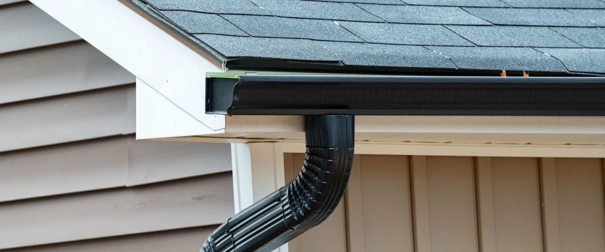 A close-up view of a newly installed black k-style rain gutter and matching downspout attached to a house with tan siding and a grey shingle roof.