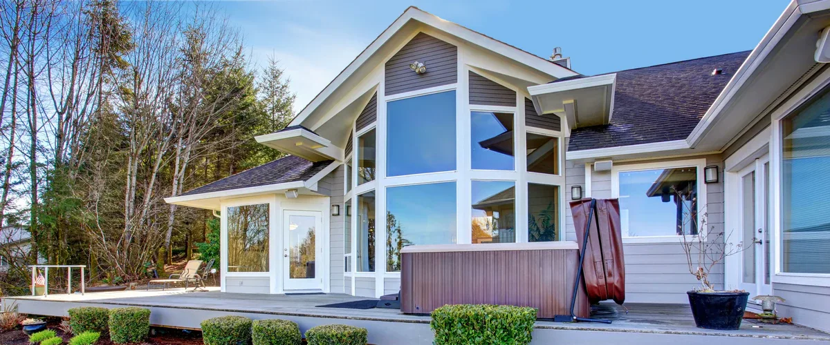 Exterior view of a contemporary luxury home featuring expansive custom-shaped glass windows and a wooden patio deck with a hot tub.