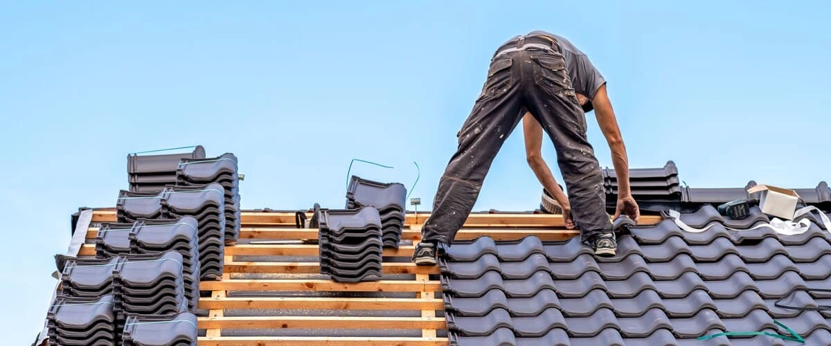 A professional roofer in work clothes carefully aligning and installing dark grey interlocking ceramic clay tiles on a wooden roof frame against a clear blue sky.
