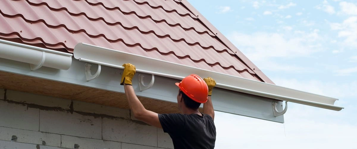 A contractor wearing an orange hard hat and yellow work gloves installs a white plastic rain gutter section onto the fascia of a house with a red metal tile roof.