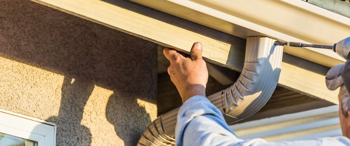 A professional contractor using a power drill to secure a white aluminum gutter downspout elbow to the side of a residential home under a roofline.