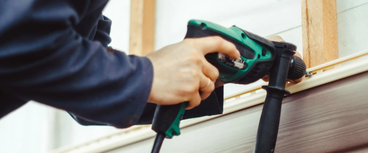 A close-up shot of a contractor's hands using a green electric power drill to secure a panel during a home siding repair project on a brown horizontal plank exterior.