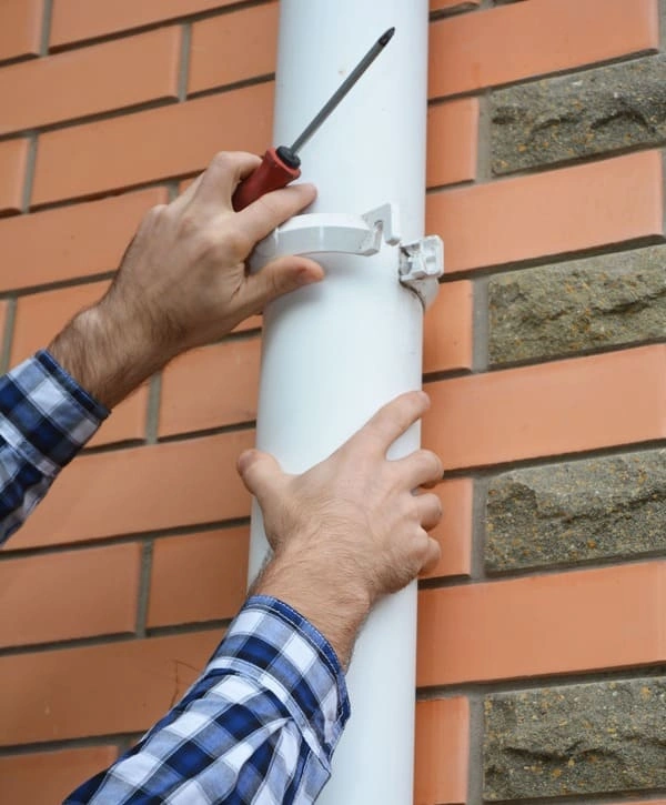 A close-up shot of a technician’s hands using a screwdriver to secure a white downspout bracket against a brick wall during a gutter repair in Holmdel, NJ.