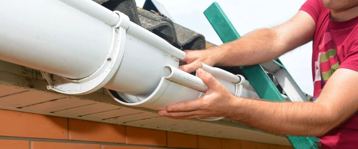 A close-up view of a person wearing a red shirt standing on a ladder while carefully installing or repairing a section of a white PVC rain gutter on a brick residential building.