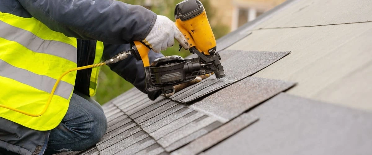 A roofing contractor in a high-visibility safety vest using a yellow pneumatic nail gun to secure asphalt shingles during a residential roof repair project.