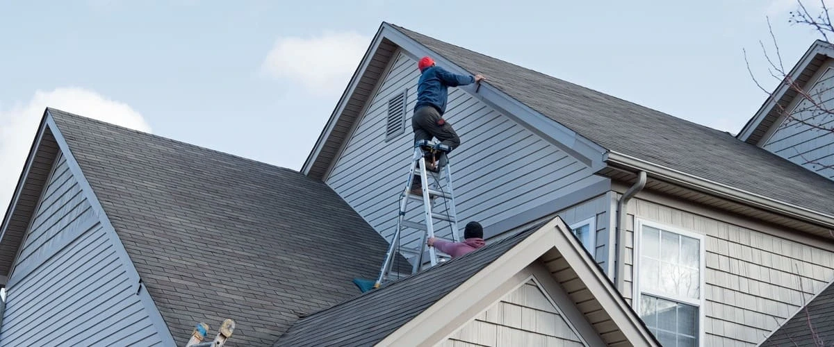Two workers using a tall silver ladder on a roof to access and perform siding repair on the upper gable of a grey multi-story suburban house.