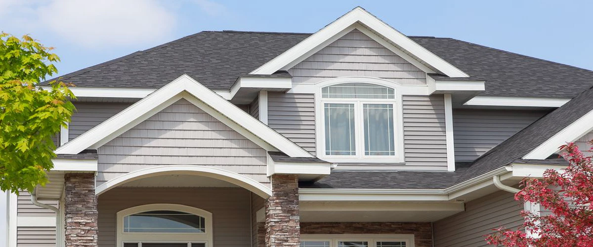 The upper exterior of a suburban house featuring light gray horizontal siding, gabled rooflines, white trim, and decorative stone pillars.