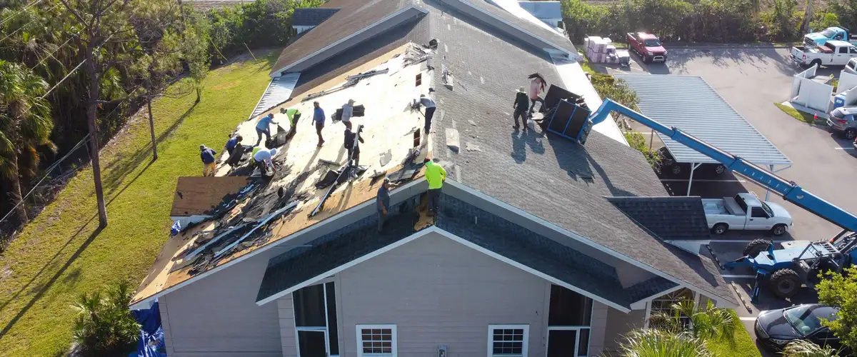 An aerial drone shot of a large roofing crew working together to repair several sections of a residential roof damaged by a storm.