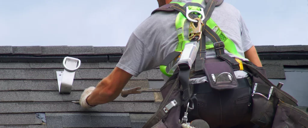 A close-up view of a roofer wearing a bright safety harness and tool belt while repairing asphalt shingles on a roof.