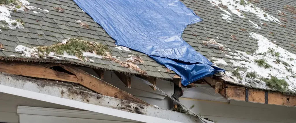 A blue emergency tarp covering a roof section damaged by ice dams and winter weather buildup.
