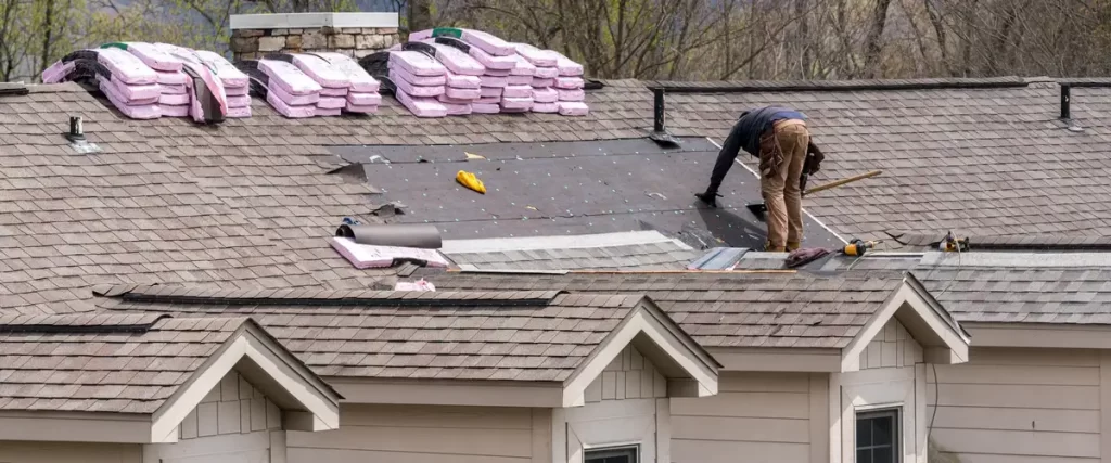 A wide shot of a house undergoing a roof replacement showing pink insulation stacks and a worker preparing the underlayment.