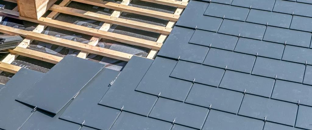 Professional installation of dark gray natural slate roofing tiles over a wooden structural frame and waterproof underlayment.