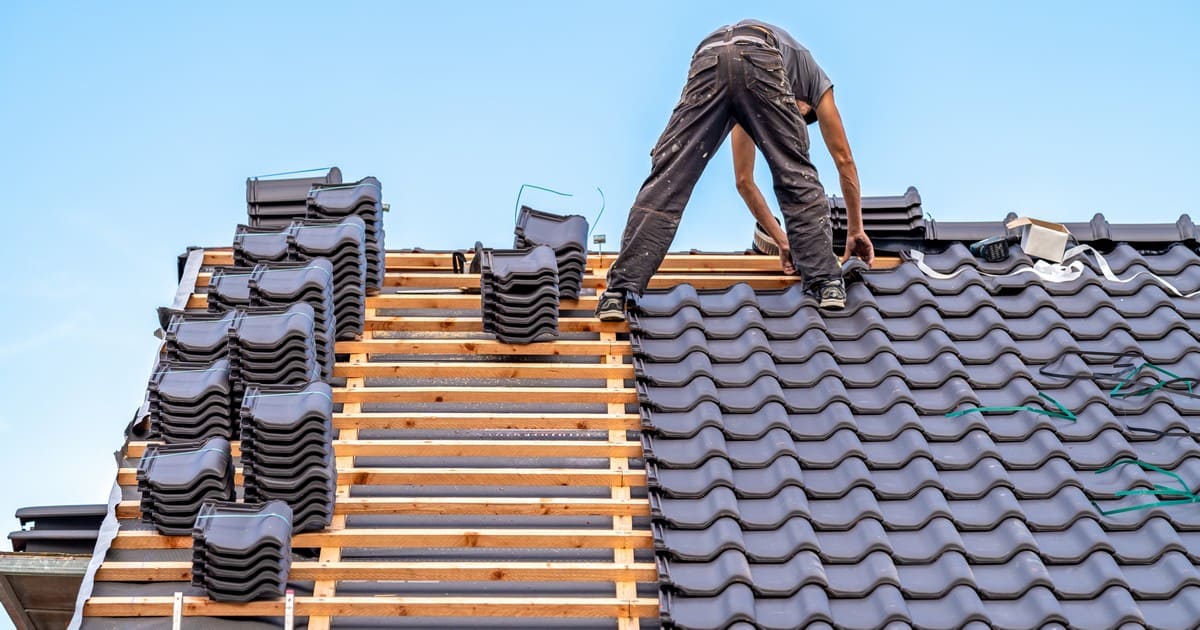 A professional roofing contractor carefully installing a new roof with dark ceramic tiles on a residential home under a clear blue sky.