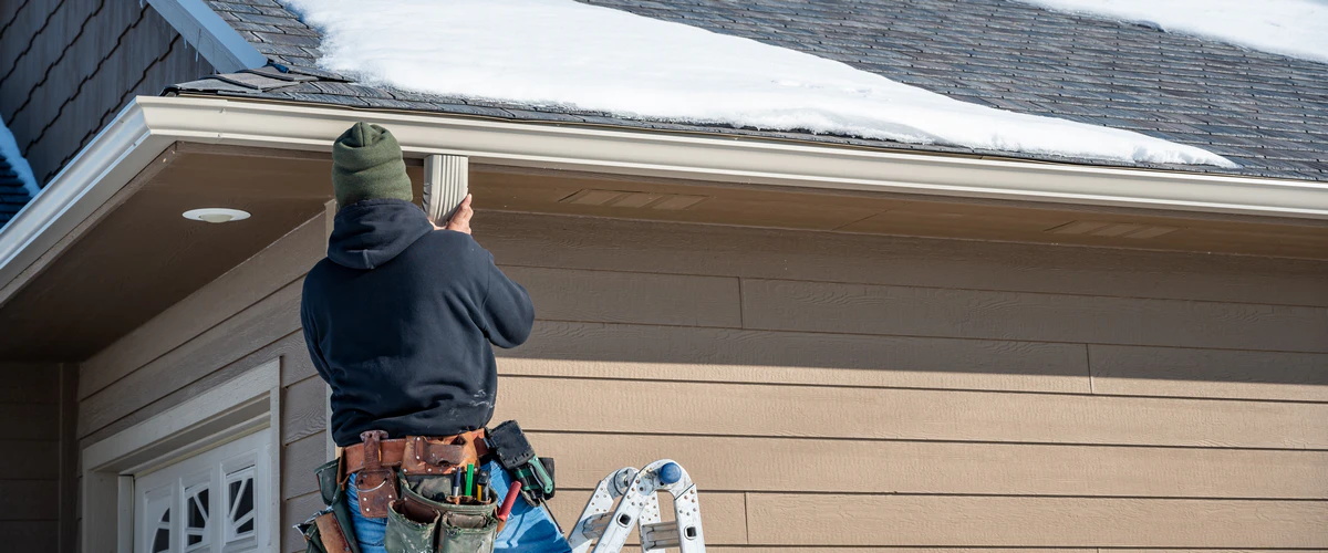 A professional contractor on a ladder inspecting and repairing a residential roof gutter system covered in snow.