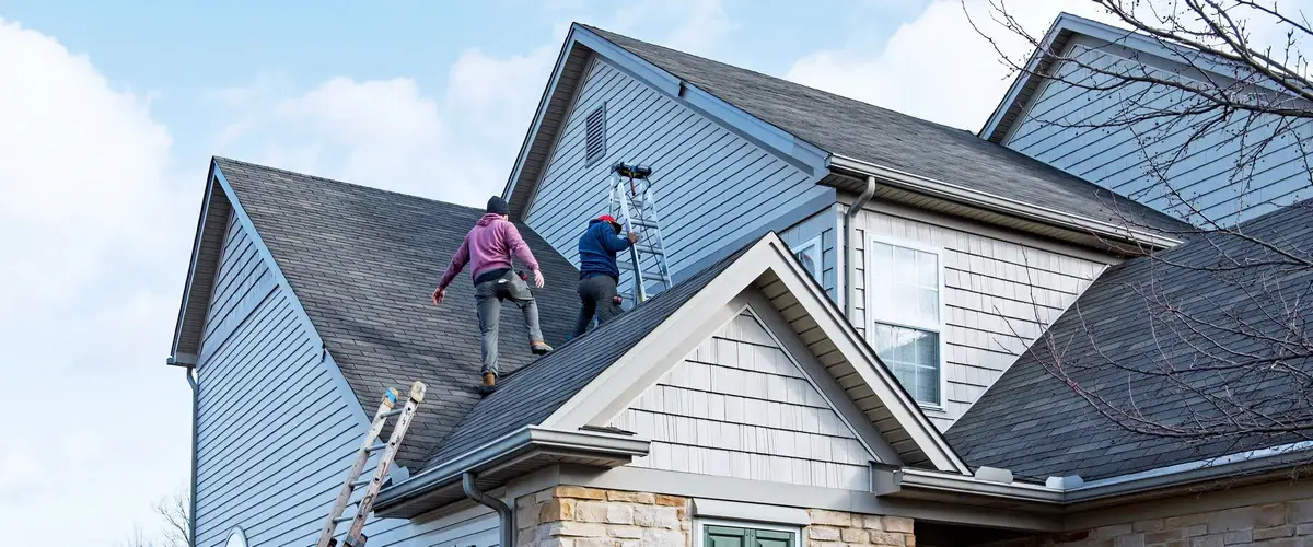 Two roofing contractors standing on a steep gabled roof using ladders to perform repair work following a wind storm.