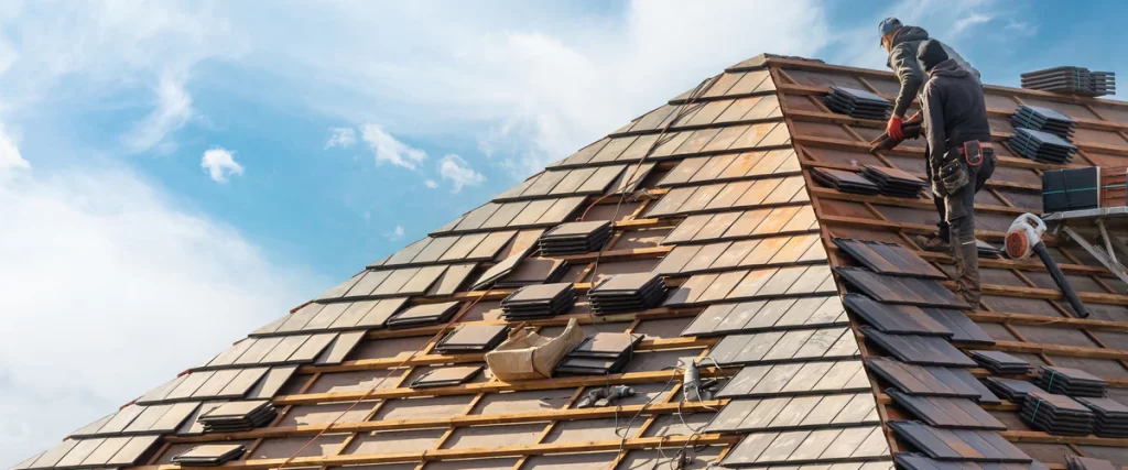 Two roofing workers collaborating to install rows of clay tiles on a steep sloped roof of a modern house.
