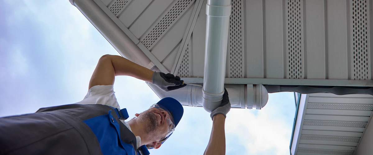 A close-up view of a technician using tools to securely install a new white aluminum gutter downspout under the roof eaves.
