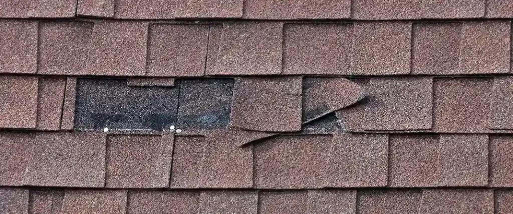 Close-up shot of reddish-brown asphalt shingles with a large gap of missing shingles, exposing the black underlayment and two silver roofing nails.