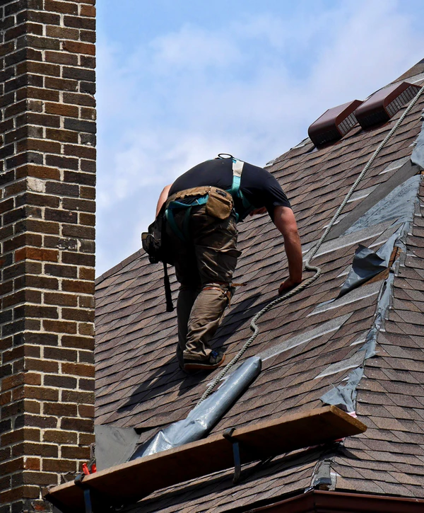 A professional roofing contractor performing emergency roof repair in New Jersey, wearing a safety harness while inspecting shingles next to a tall brick chimney.