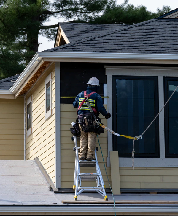 A professional contractor in a safety harness on a ladder measuring a yellow house exterior for an emergency siding repair in New Jersey.