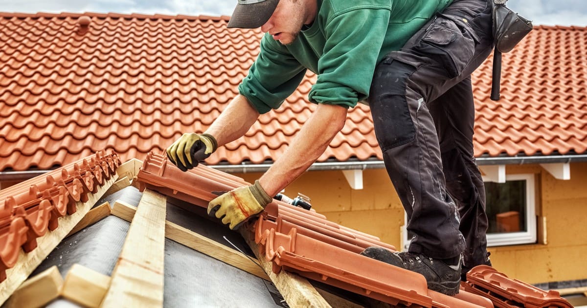 A professional roofing contractor wearing gloves and a green shirt carefully laying red terracotta clay tiles on a residential roof.