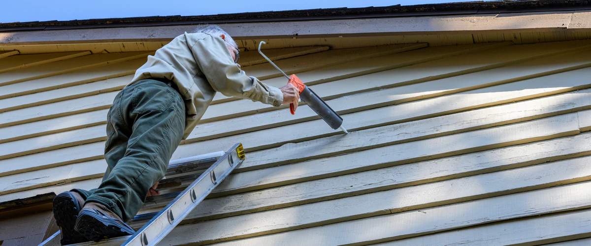 An experienced worker on a ladder using a caulking gun to apply sealant to the seams of horizontal siding during a siding repair service.