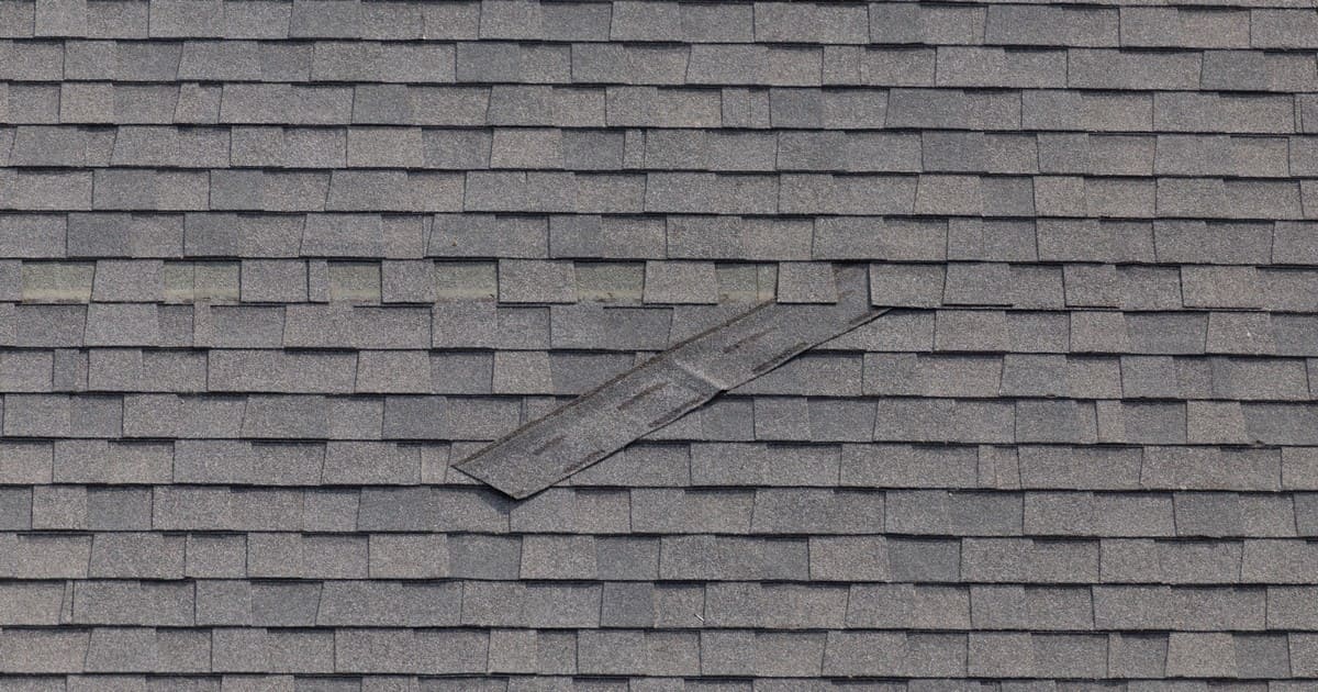 A wide view of a grey architectural shingle roof showing a loose, detached shingle sliding out of place, resulting in missing shingles on roof.