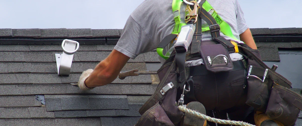 Close-up of a roofing professional in a safety harness performing roof repair by using a pry tool to adjust asphalt shingles near a permanent roof anchor.