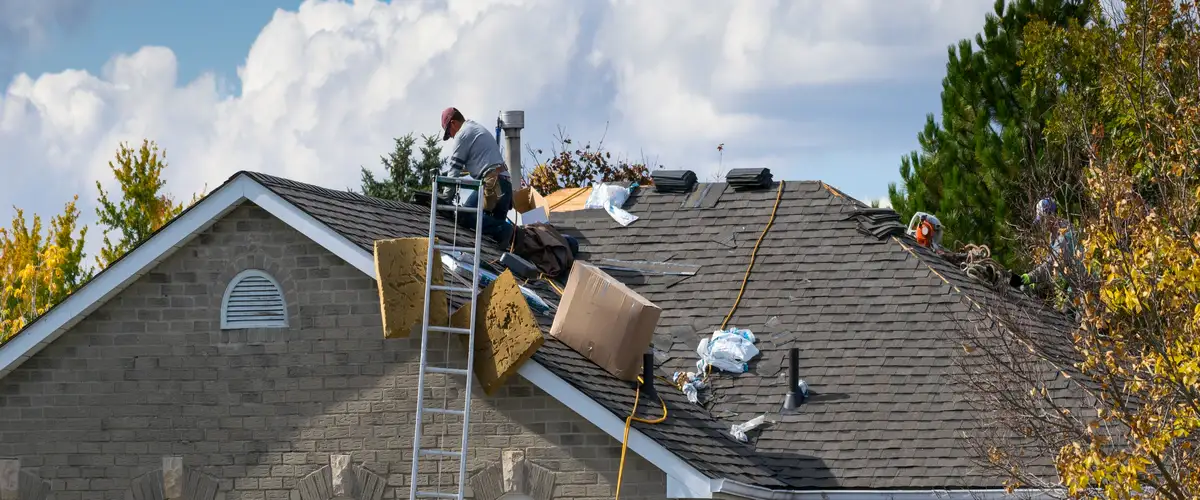 A wide view of a residential roof repair project in progress, showing contractors, bundles of new shingles, and a tall ladder leaning against a brick home.
