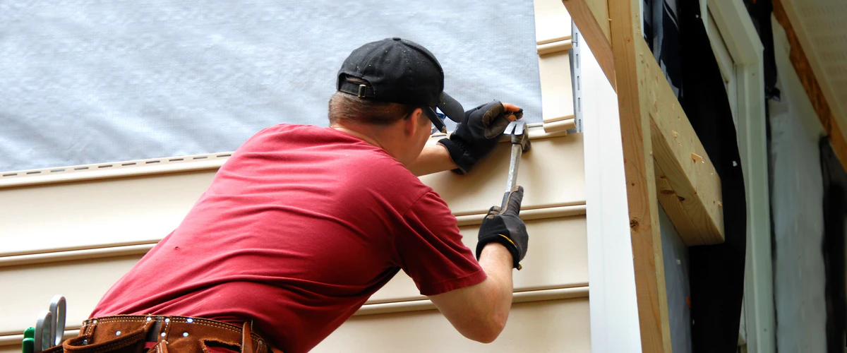 Close-up of a technician in a red shirt and black cap using a hammer to nail tan vinyl siding panels during a siding repair project.