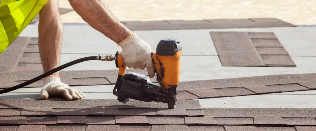 Close-up view of a roofing contractor's gloved hand using an orange pneumatic nailer to secure architectural asphalt shingles.