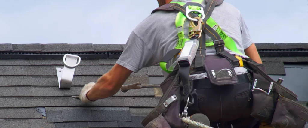 Back view of a roofing contractor wearing a lime green safety harness and fall protection equipment while working on dark asphalt shingles.