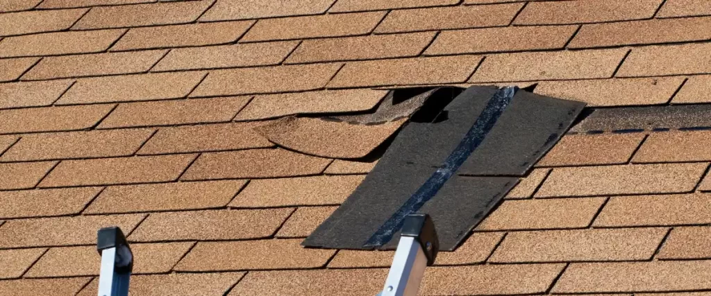 High-angle view of tan-colored shingles with a section of missing shingles and a ladder visible at the bottom, revealing the black sealant strip beneath.