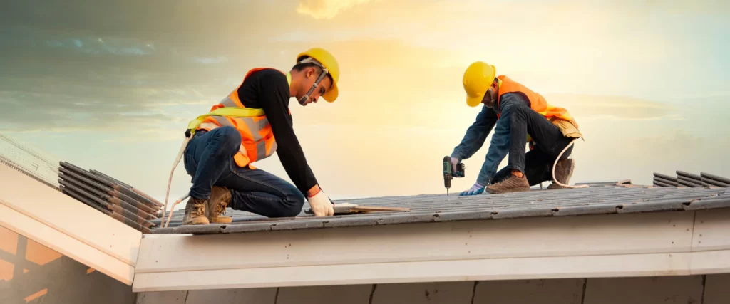 Two roofing contractors wearing yellow hard hats and safety vests installing grey roof tiles against a sunset sky background.