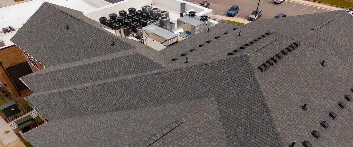 Close-up drone view of a complex grey ashpalt shingle roof showing intersecting valleys, ridges, roof vents, and an adjacent flat roof section housing commercial HVAC units.