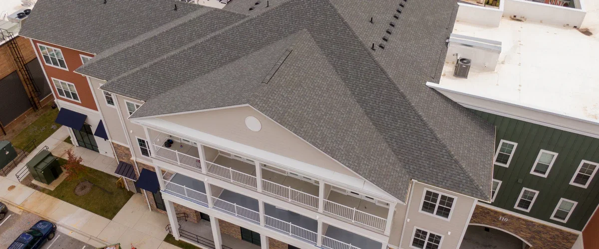 Exterior view of a multi-story residential building highlighting covered white balconies, brick accents, and a large gable end covered by a grey ashpalt shingle roof.