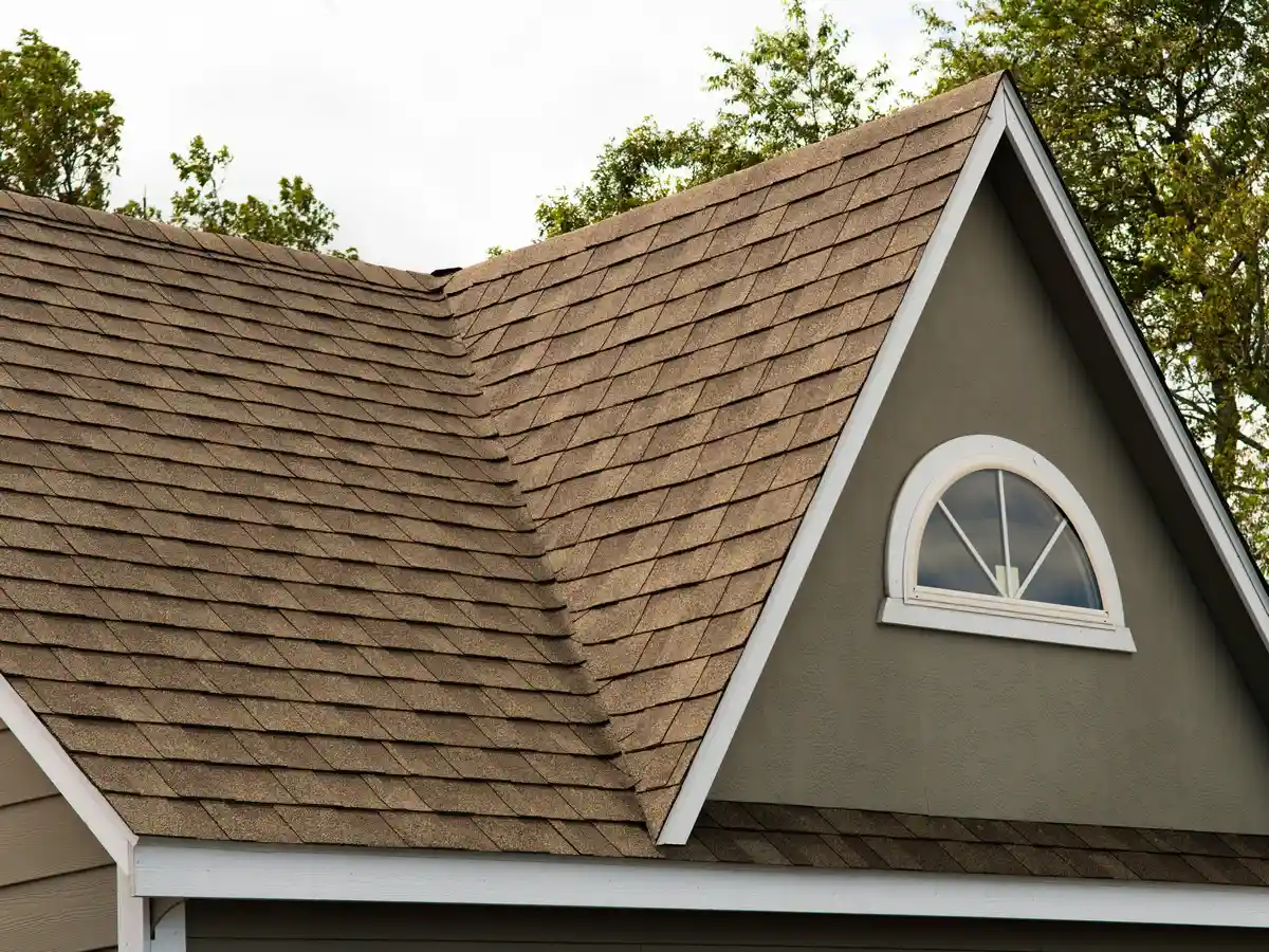 A steep gable roof covered in dimensional brown asphalt shingles, featuring a white arched half-round window on a tan house exterior.