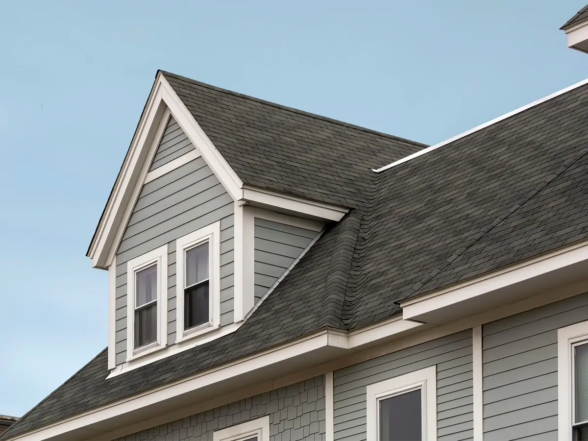 A residential home featuring a dark grey asphalt shingle roof and a front dormer with two white-framed windows against a clear blue sky.