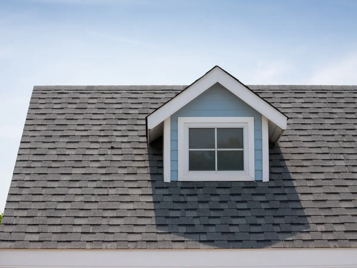 Close-up of a dark grey asphalt shingle roof featuring a small light blue dormer with a single white-framed, four-pane window under a bright blue sky.