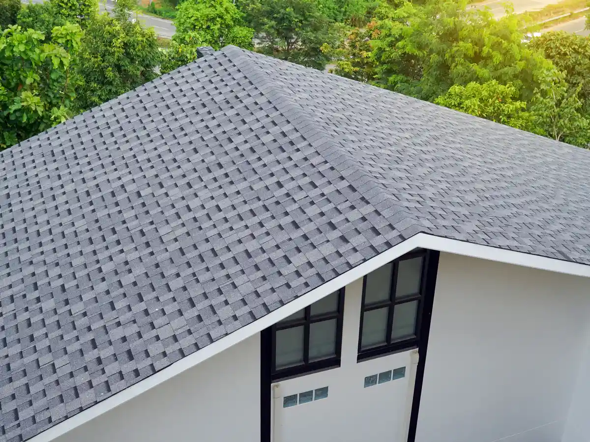 Aerial view of a modern white house showcasing a unique woven or checkerboard pattern grey asphalt shingle roof, with lush green trees in the background.